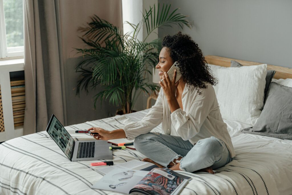 A woman works from home, multitasking on her laptop and phone while sitting on her bed.